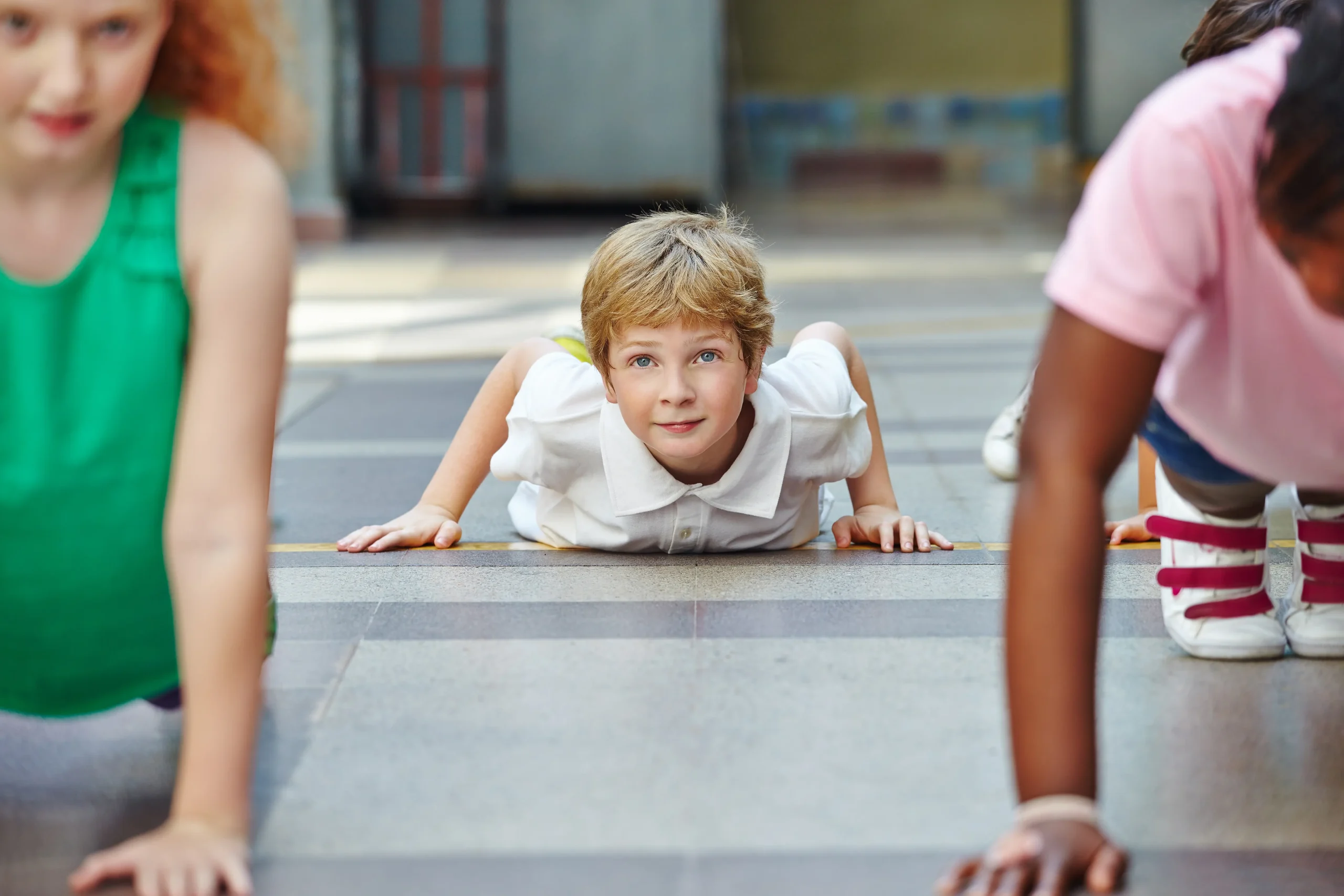 Three children performing push-ups on a gym floor, with a young boy in a white shirt smiling at the camera.